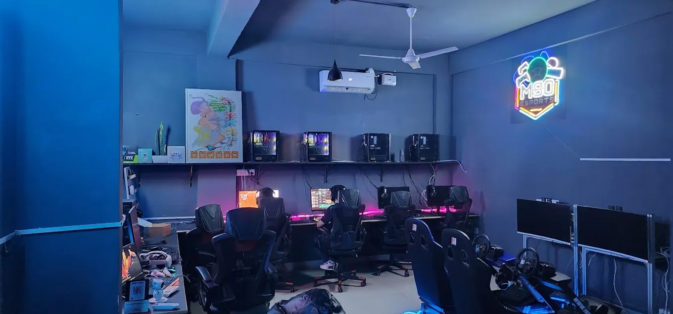 Inside the M80 Esports café. A row of gaming PCs on pink LED-lit desks with players seen from behind, two racing-simulator rigs with steering wheels and bucket seats on the right, and the M80 Esports neon sign on the wall.