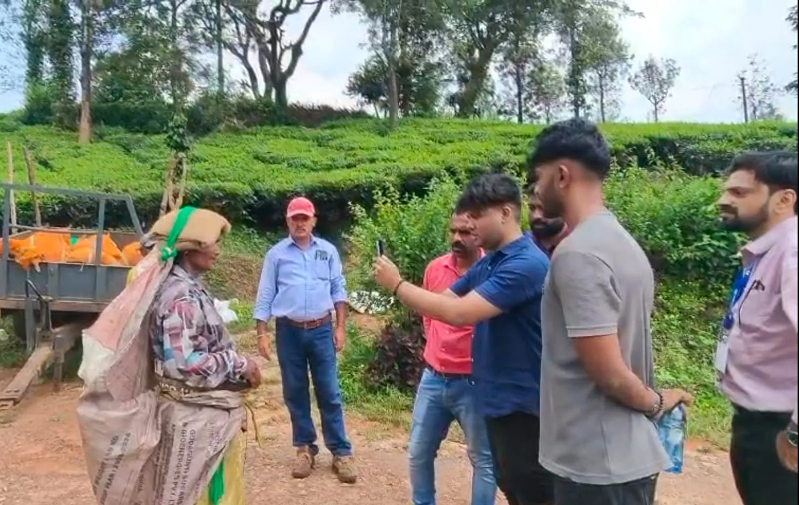 On a tea estate, one of our team holds up a phone in front of a tea picker carrying a sack of leaves, while estate staff and the rest of us watch.